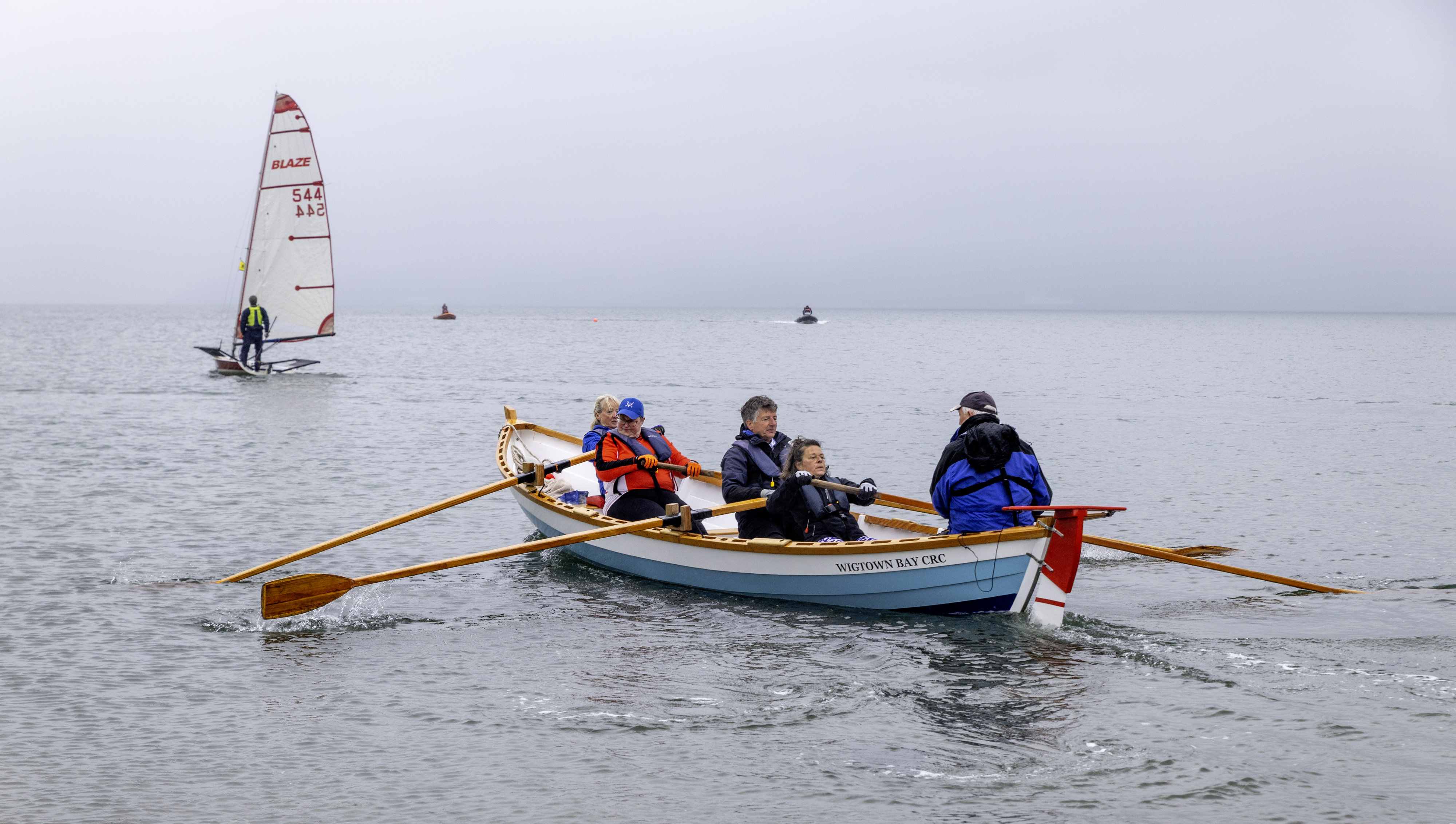 A coastal rowing boat in the foreground with a sailing boat in the background