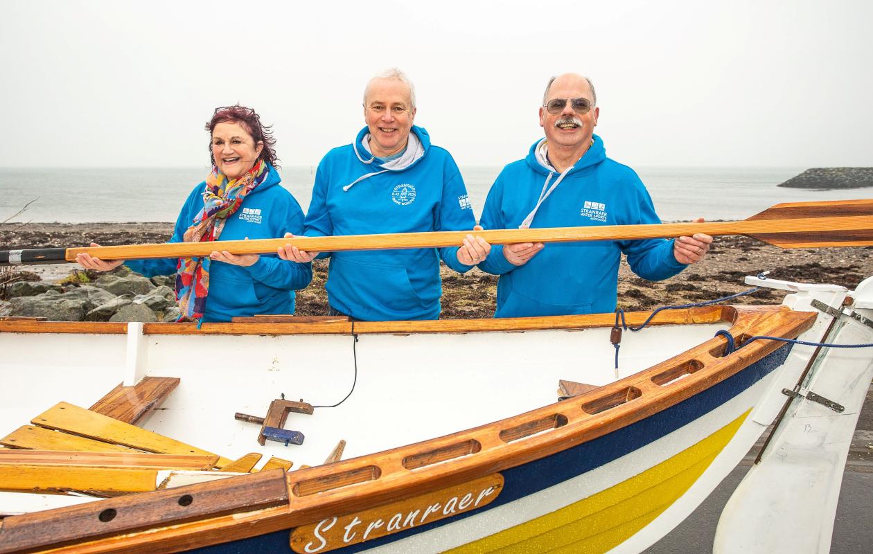 Three people holding an oar alongside a coastal rowing boat